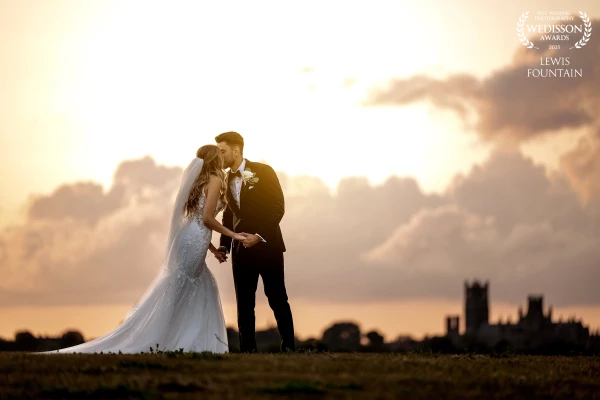 Fran and Elliot sharing a tender kiss with the majestic Ely Cathedral in the distance, under a dramatic golden sky.