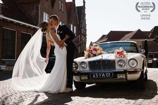 “A radiant bride and groom beside a charming old timer, flowers bursting with color, their kiss says...