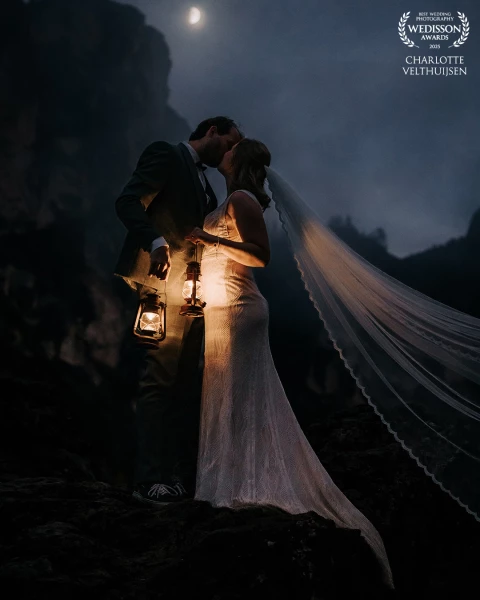 Beneath a sliver of moon and the hush of the Dolomites, lantern glow warms their kiss, an intimate,...