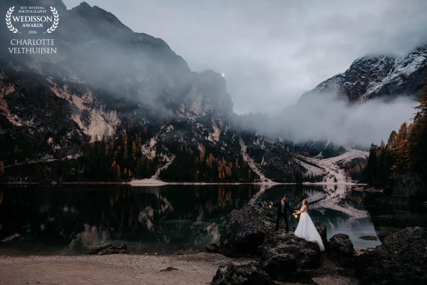 On the edge of a glassy Dolomite lake, they reach for each other with lanterns glowing softly in the...
