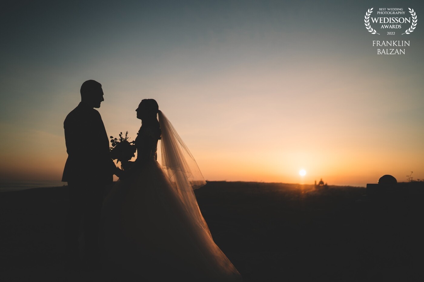 The photograph features a couple who were married on the island of Gozo, during a beautiful sunset. The setting and lighting create a romantic and striking image, with the couple's silhouettes standing out against the glowing sunset in the background. The photograph may have been taken just after the wedding ceremony, adding to the sentimental value of the image.