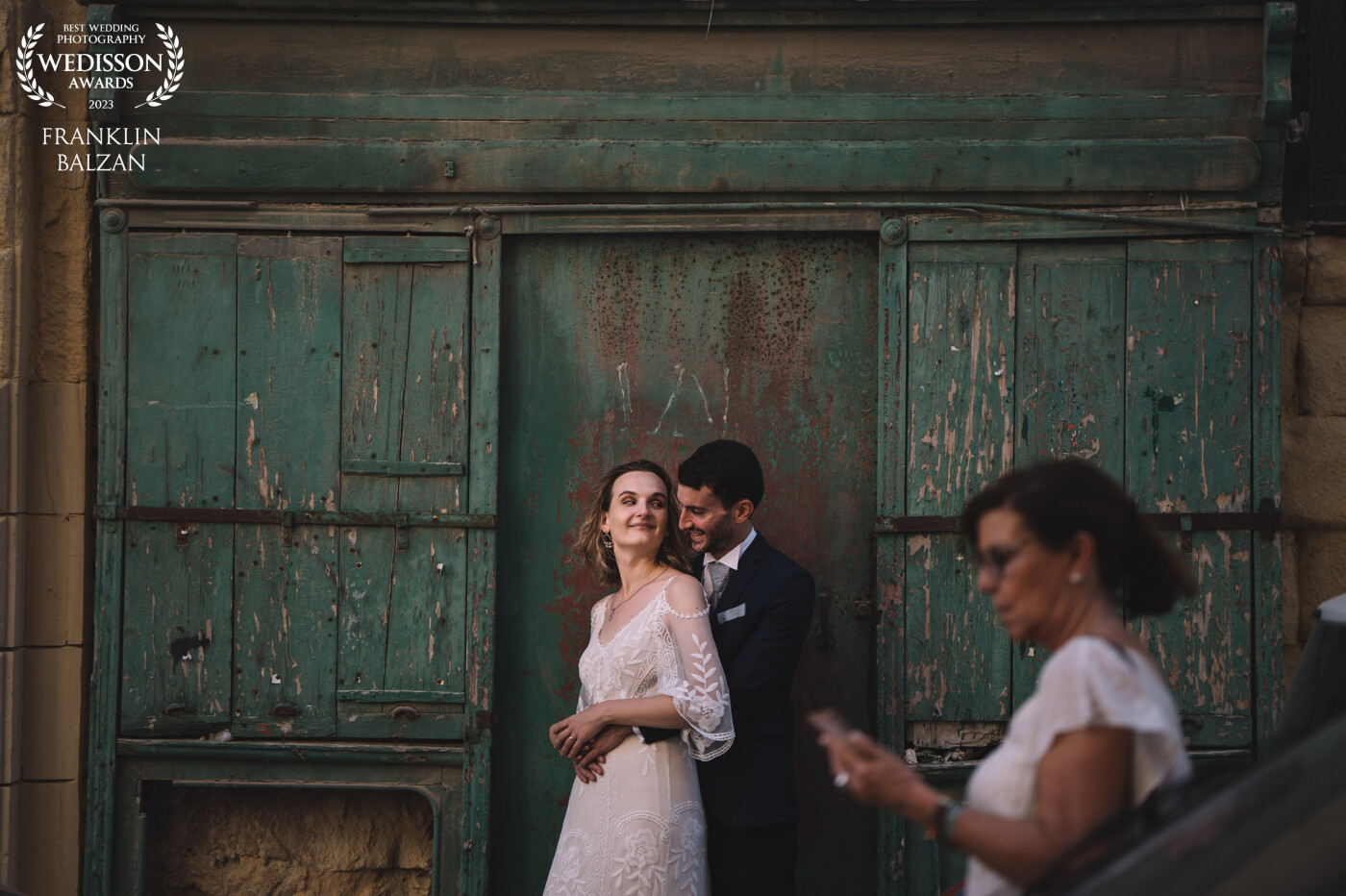 The wedding image is set against the backdrop of an old wooden green door in the heart of Valletta, Malta's capital city. The couple stands in front of the door, their bodies touching and in contact as they glance at each other.  A blur of motion in the foreground reveals a woman tourist passing by, adding a touch of local flavor and authenticity to the image.