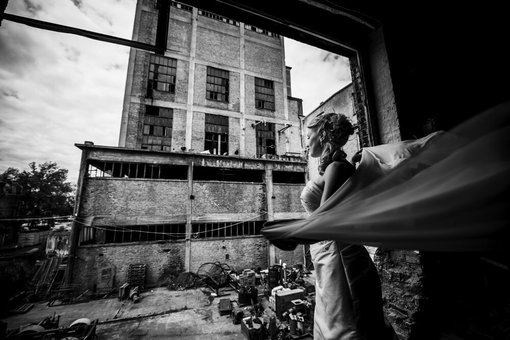 This picture was taken at an abandoned paper factory. There you have the ruins in contrast to the beautiful bride. Inside a building the whole wall was missing, so the bride went near to the edge and at the right moment the wind caught her bridal veil.