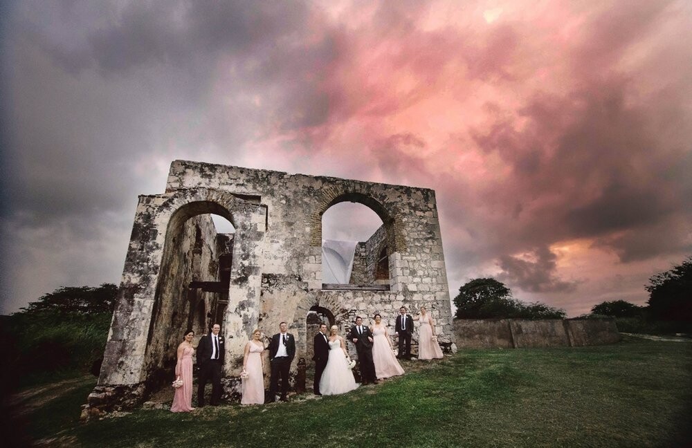 Location: Montego Bay, Jamaica.<br />
Ashley and Anton are avid golfers and accordingly they wanted to incorporate their passion for the sport when choosing the venue for their destination wedding. They found the perfect location at the Hilton Rose Resort and Spa in Montego Bay, Jamaica at the historic 18th century aqueduct ruins overlooking the greens of the Cinnamon Hill Golf Club. These aqueducts were originally used to irrigate sugar cane plantations, bringing fresh water down to the crops from the hills above. Their wedding ceremony took place as the sun was setting over the lush green hills of Montego Bay. This photo was captured just after the sun had set, leaving the sky full of beautiful pink and grey tones.