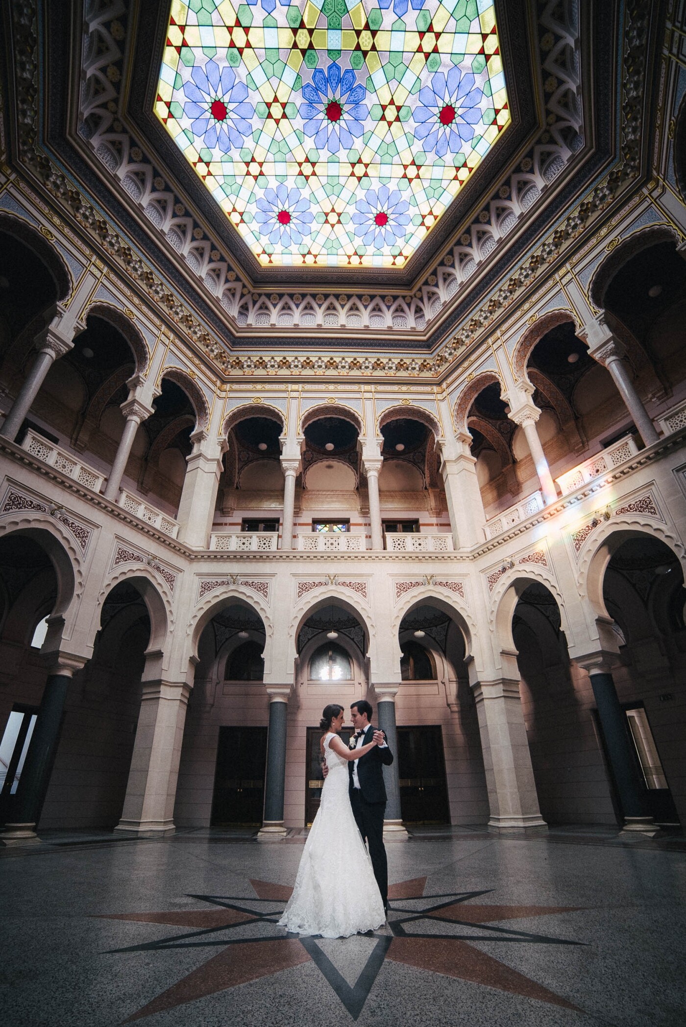 The photo was taken in the Sarajevo City Hall, an old neo-moorish architecture building. We were lucky to be allowed to have a photosession there after closing time, so it was empty, as soon as we entered I had the idea to show the architecture around the bride and groom dancing. So I used my 14mm lens on a full frame camera to get the perspective and the wide shot. I needed an ultra-wide lens to catch them dancing as well as the colorful glass roof.