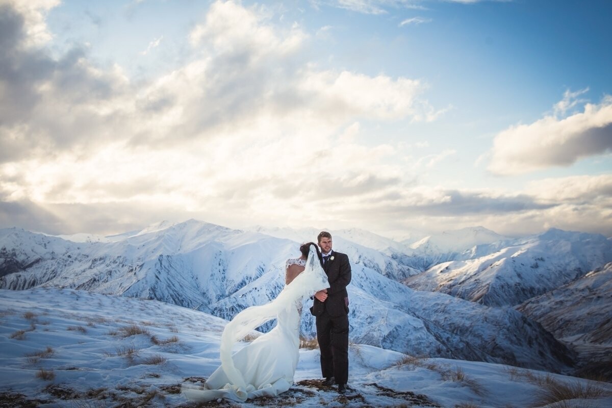 It was a bitter winter’s day when David and Jacqui tied the knot in Queenstown, New Zealand. <br />
They flew over with their entire families from Sydney, Australia to have their wedding in the mountains, and wanted their couple shoot up on the snow. <br />
We choppered up onto a nearby mountain top, and there was a freezing wind blowing. <br />
I found the best spot looking back over the mountains in the low afternoon light, and posed them with Jacqui’s back to me as I wanted to show off the beautiful low cut of her Steven Khalil dress and veil flying in the wind.<br />
I also wanted her leaning into David to symbolise the surrender and safety of marriage. <br />
At one point I thought Jacqui was going to get hypothermia! But she was incredibly resilient and endured the deep cold to take a collection of stunning photos, including this one. 