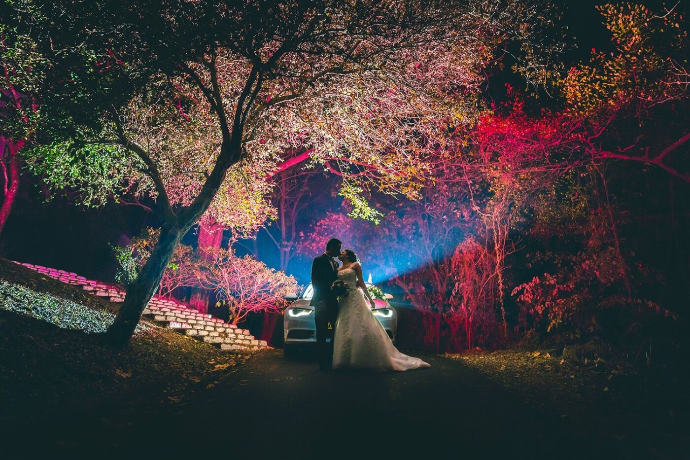 We were done with the couple shoot of Charlston & Fomina and were headed back to their car to head to the reception, When i thought it would be great to try out a shot with the Audi in the background, using its lights and my flash triggered from behind. What we did not expect was the rear brae lights of the car to create these crazy colors on the trees behind the car, which formed such a beautiful colorful pic.<br />
Turned out to be very magical as the couple shared an intimate moment.
