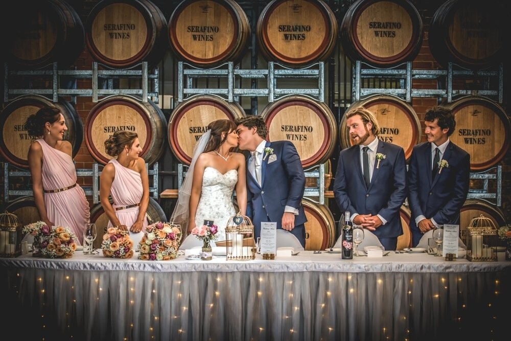 We love winery weddings & what I love above Serafino Wines in McLaren Vale is the bridal table in front of the big stack of wine barrels. Not only is it a great backdrop, it helps set the scene for a wedding in one of the best wine regions in Australia. How adorable is Sara & Josh kissing, as their bridal party look on? This photo was taken during a quick preview of the room before the guests arrived. It's always a good idea for the bride & groom to have a look at the reception before the guests arrive. As sometimes this is the only time they get to have a look at the room fully decorated for their own wedding. It will never look the same once the guests arrive.