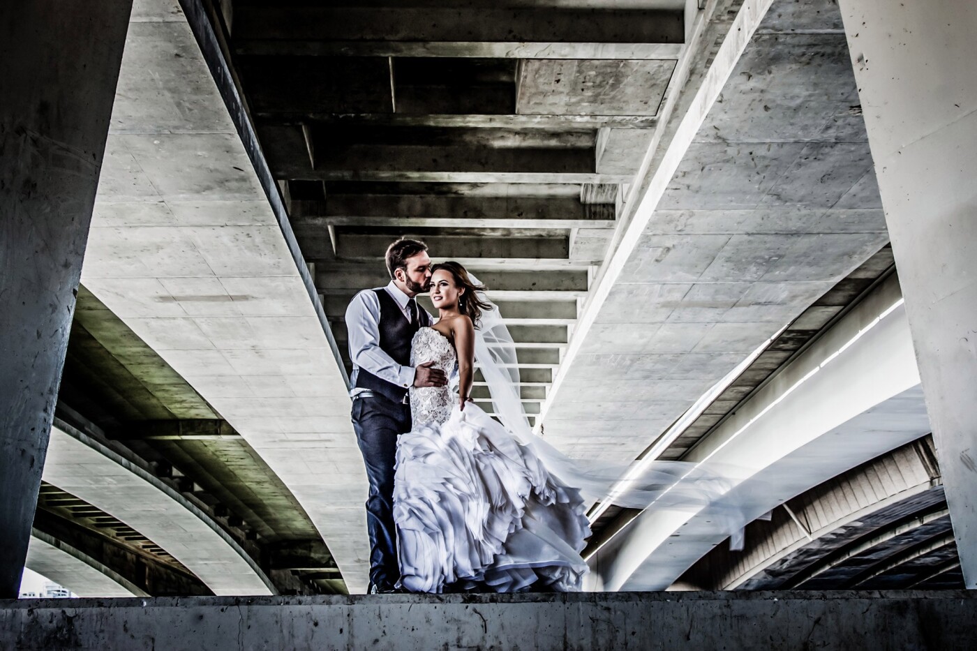 This image was captured underneath a bridge spanning the Swan River in Perth. Our couple wanted a shot across the river showing the city skyline. Whilst we were there we kept looking at the strong lines of the underneath part of the bridge. So, we lay on the floor to see if it would work. It did, so we got our bride and groom up on the concrete wall at the river’s edge. We waited for the wind to come and ruffle the layers of her amazing, couture wedding gown and her veil and create this shot. We just love it. Our bride wanted images fit for a magazine; and that is what we gave her. We are very proud of the fact that all our wedding images submitted in competitions are of real brides and grooms on their actual wedding day.
