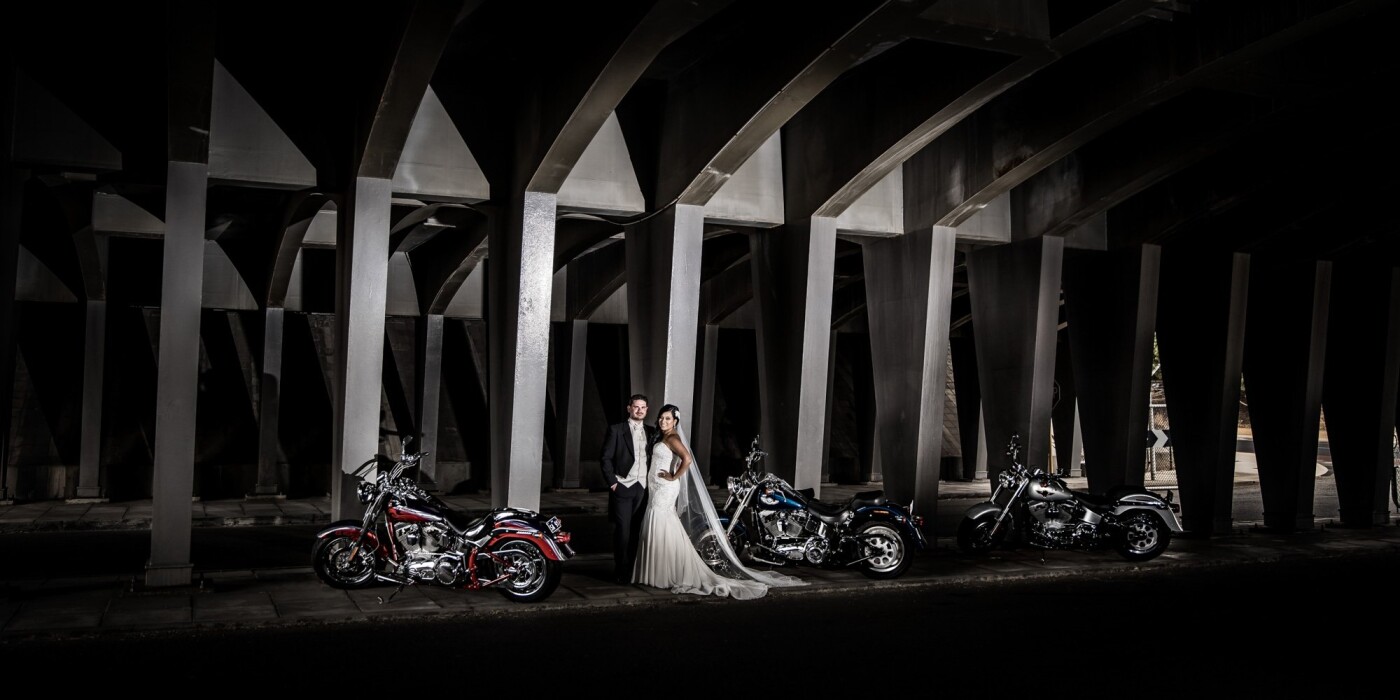 Our couple, Yaira + Sean had their wedding in early January on a super hot day, The heat was intense and there was no breeze to lessen the temperature effect. This did not stop our couple from enjoying every single minute of their wedding day though. The groom’s father brought 3 brand new Harley Davidson motorbikes especially for the wedding day, they were driven as an escort ahead of the wedding cars. So, we had to come up with a great photo idea to incorporate them in. <br />
<br />
The entire wedding party was in love with the idea of doing a shot under this old bridge in Fremantle, WA. They got some much needed shade from the sun’s intense heat; and the low light + concrete walls and columns were a great background to create this shot.<br />
<br />
We used 4 off camera flashes to illuminate the couple and the bikes so that we could darken everything else right down.<br />
<br />
This image is now a double page spread in their wedding album.