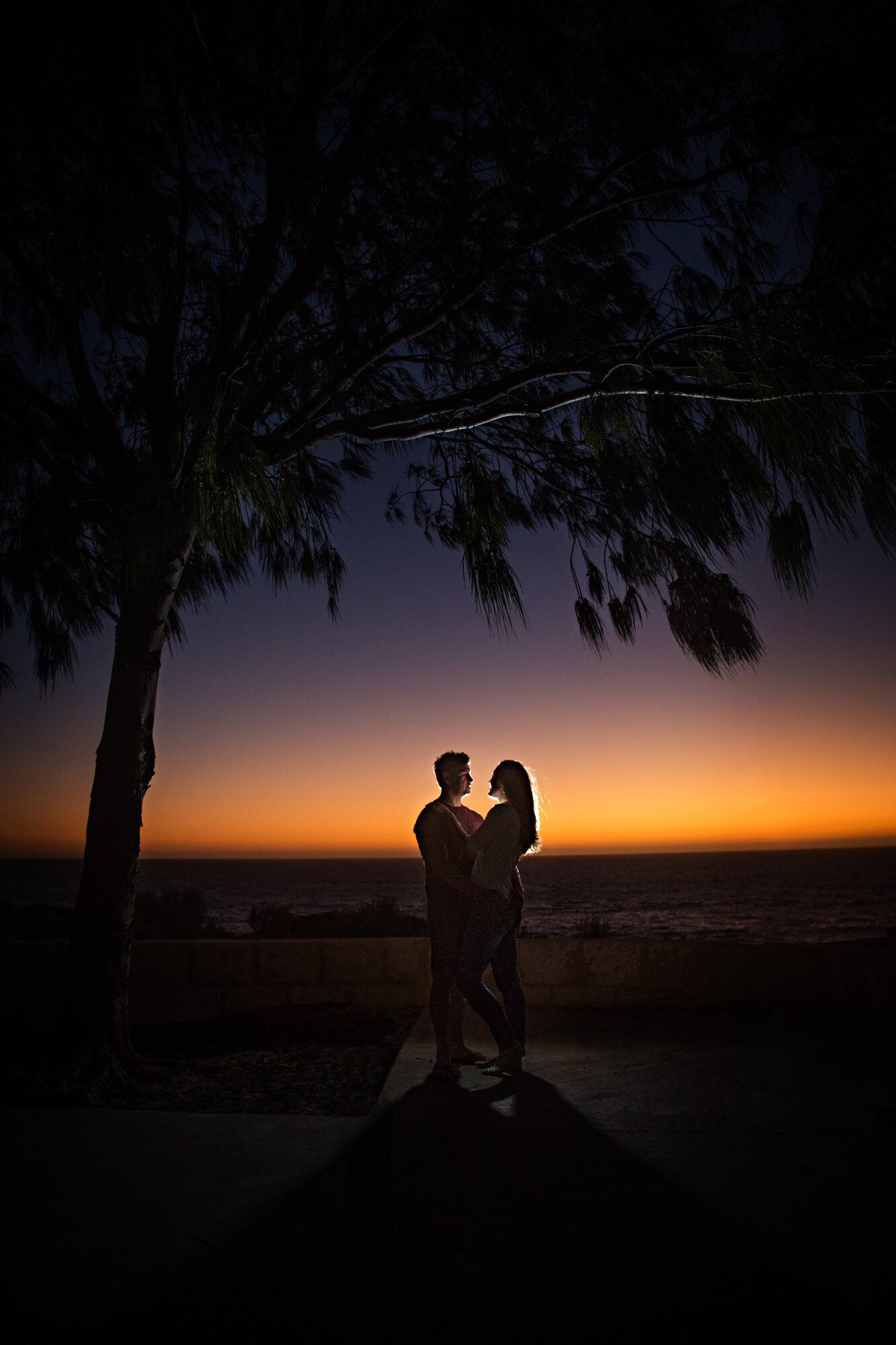 We were photographing Sarah + Mark for their pre wedding photo shoot. They asked if we could head to their favourite beach. It was a gorgeous afternoon, but the sky was pristine clear. Our couple were hoping for some clouds and a beautiful sunset. The sunset wasn’t too special so after a couple of shots we headed back to the car park. When we turned around there was an amazing glow in the sky, we quickly set up a couple of flashes on the ground behind our couple for some backlighting, had them stand under a little tree and captured this image. Our couple were so happy with this last shot we captured - and, so were we!