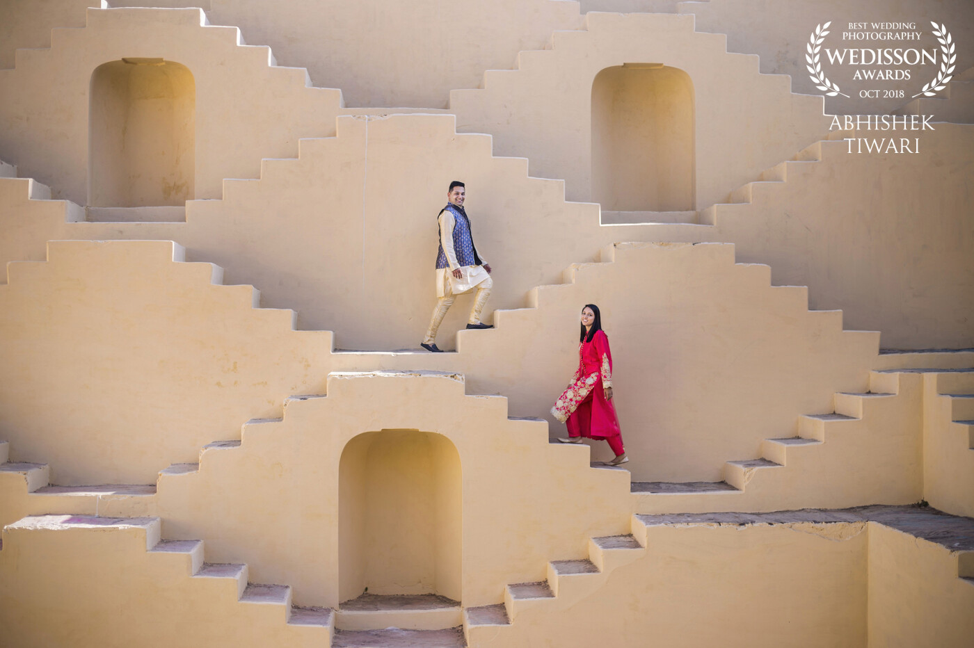 This shot was taken on the geometric staircases of the Panna Meena Ka Kund -  a sixteenth century step-well located on Jaipur. In the picture Ridhish is wearing a traditional Indian attire and walking up on the higher stairs of the eight storey stepwell while Chandni, dressed in a red traditional suit is on the lower stairs. Although they're both standing apart, their body movements are coordinated and they are facing the camera and smiling. This aspect along with the criss-cross stair patterns, that although situated away have a certainty of arrangement within, sets this picture apart from other traditional wedding photographs where intimacy between the couple is encouraged. 