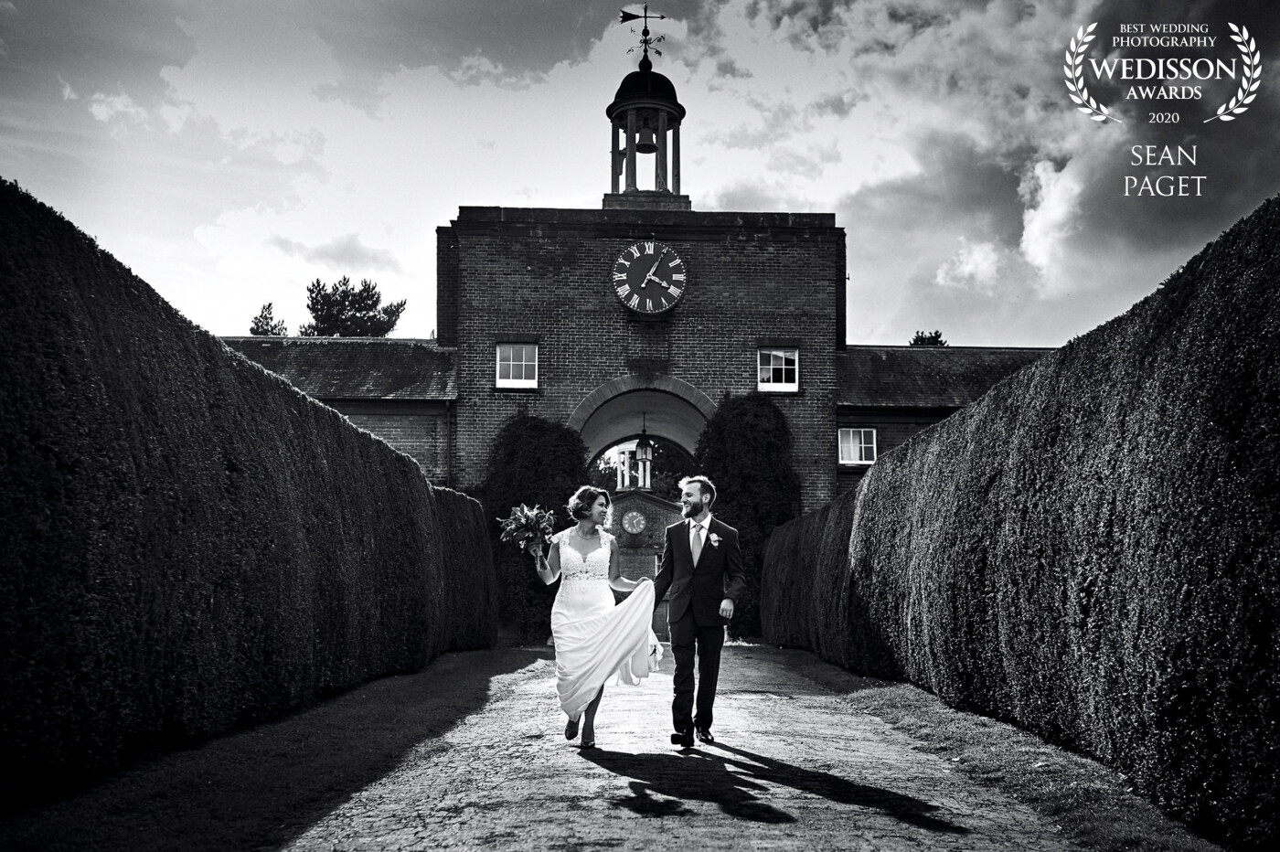 We'd just come back from a walk around the large estate at Walcot Hall in Shropshire, UK, shooting some bride and groom portraits. On the way back to the Hall for the wedding breakfast, I snapped a few shots walking backward on the way and this fell out. I just love the shadows and the symmetry that the hedges and the clock arch provided. Link to blog: https://seanpaget.com/eclectic-walcot-hall-wedding-photography/