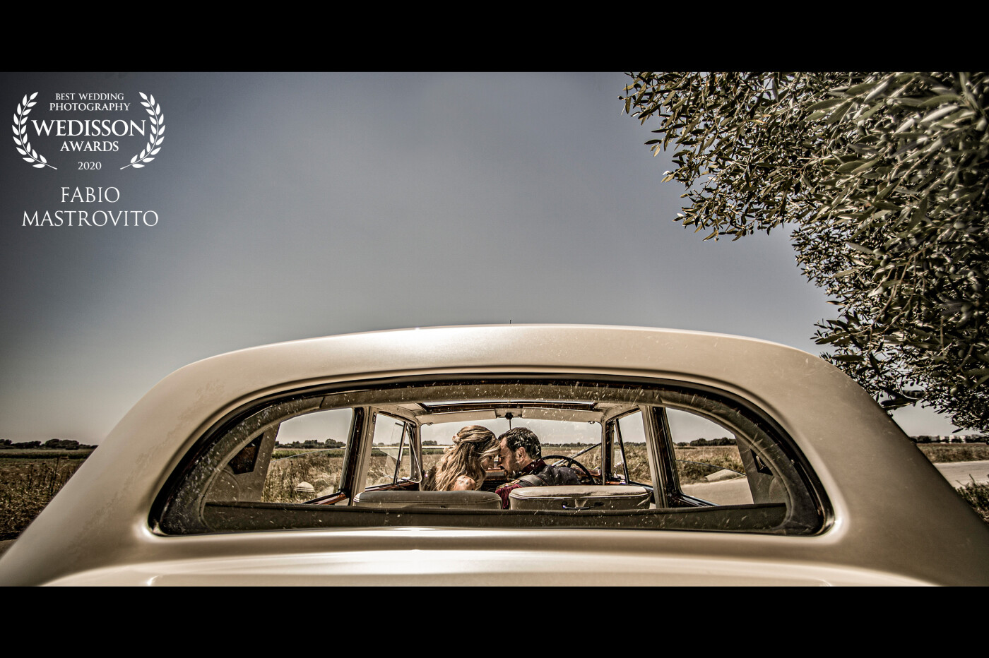 Scatto grandangolare, coppia di sposi fotografati attraverso il lunotto della loro auto vintage. Crea una prospettiva insolita, quasi surreale.