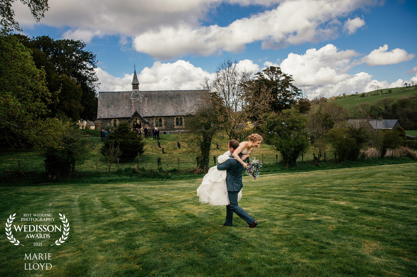 A very candid moment. The Brides shoes were sinking into the grass, so her Groom offered her some help. The image features their church in the background.