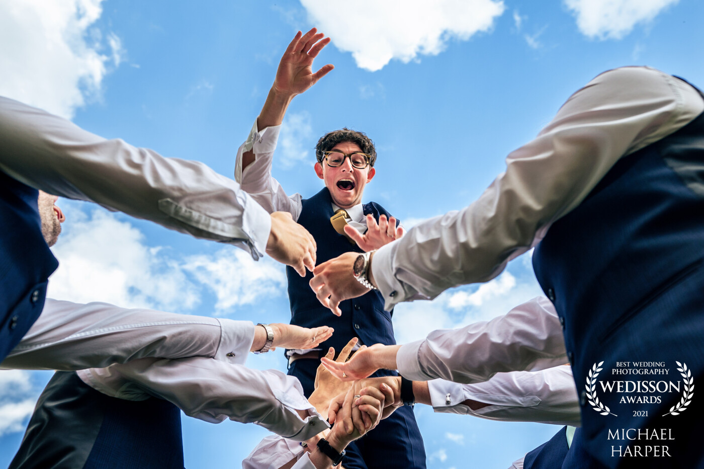 The groom wanted to do something fun and different for his groomsmen photo. After building up the courage the groom was thrown in the air by his ushers and best man. To capture this image I decided to lay on the floor and shoot up through the arms as he got air.