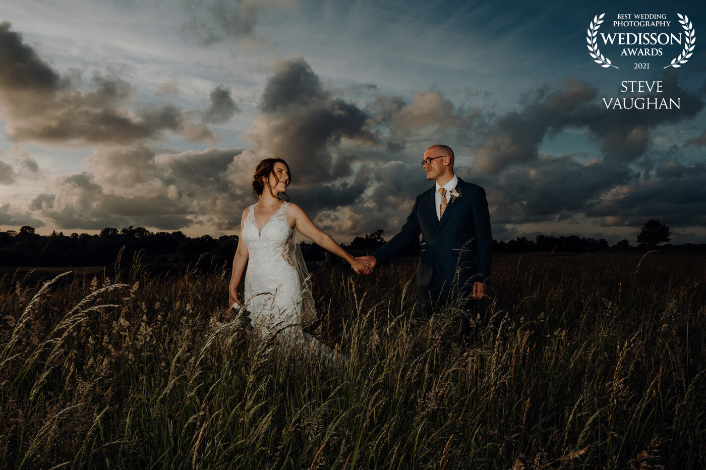 Lisa and Rob walking through the long grass in the late spring evening after their wedding at Dovecote Barn and Events, Adderbury, Oxfordshire UK.