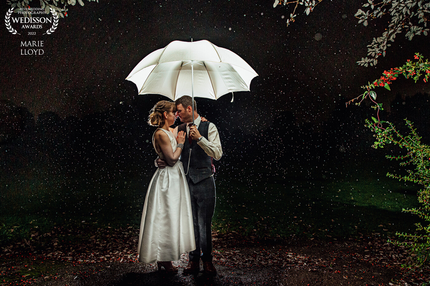 The couple was posed under an umbrella in the dark and lit with a Speedlight from behind pointing up at the umbrella, this, in turn, lit up the rain. The foliage was used as a frame