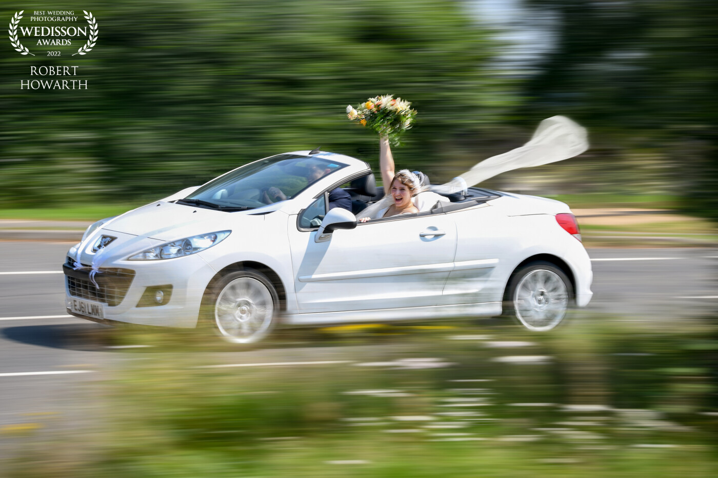 This picture was taken at a wedding in Essex, England. The bride and groom had just left the church and were driving to their reception. I asked the bride to hold her bouquet up as she went past. I used a slow shutter speed 1/20 sec to create a sense of movement as they went past.