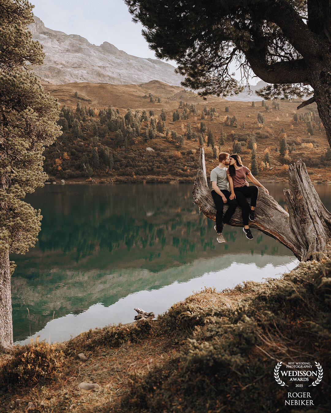 This image captures an intimate moment between two people — nestled in the breathtaking scenery of the Bernese Oberland in Switzerland. Taken during an engagement session, it speaks of serenity, connection, and the feeling of being exactly where you’re meant to be.<br />
<br />
The couple sits on a striking, weathered tree trunk above a deep emerald mountain lake — a natural throne, framed by majestic peaks and autumn-colored forest. The lake’s calm surface mirrors the landscape, adding depth and a painterly stillness to the scene.<br />
<br />
The composition feels both powerful and gentle — a perfect balance of nature’s raw beauty and quiet tenderness. This image resonates not only visually, but emotionally. It stands as a symbol of true closeness, grounded romance, and the beauty of the moment — far from anything staged.