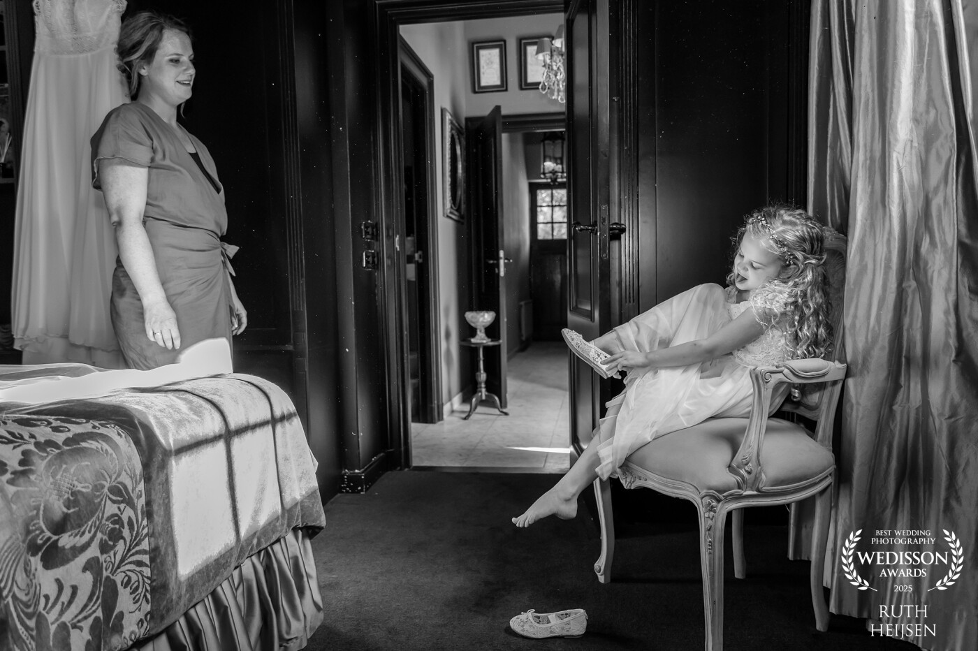 A spellbinding moment filled with pure pride and love, as the bride-to-be gazes at her daughter. With emotion in her eyes, the bride watches as her little girl (her bridesmaid) prepares with utmost focus. Dressed in a beautiful gown, the girl sits gracefully on an elegant chair, carefully slipping on her dainty shoes. It’s a tender scene that bridges generations. A mother, on her special day, completely captivated by the beauty of her child and the deep love that connects them.