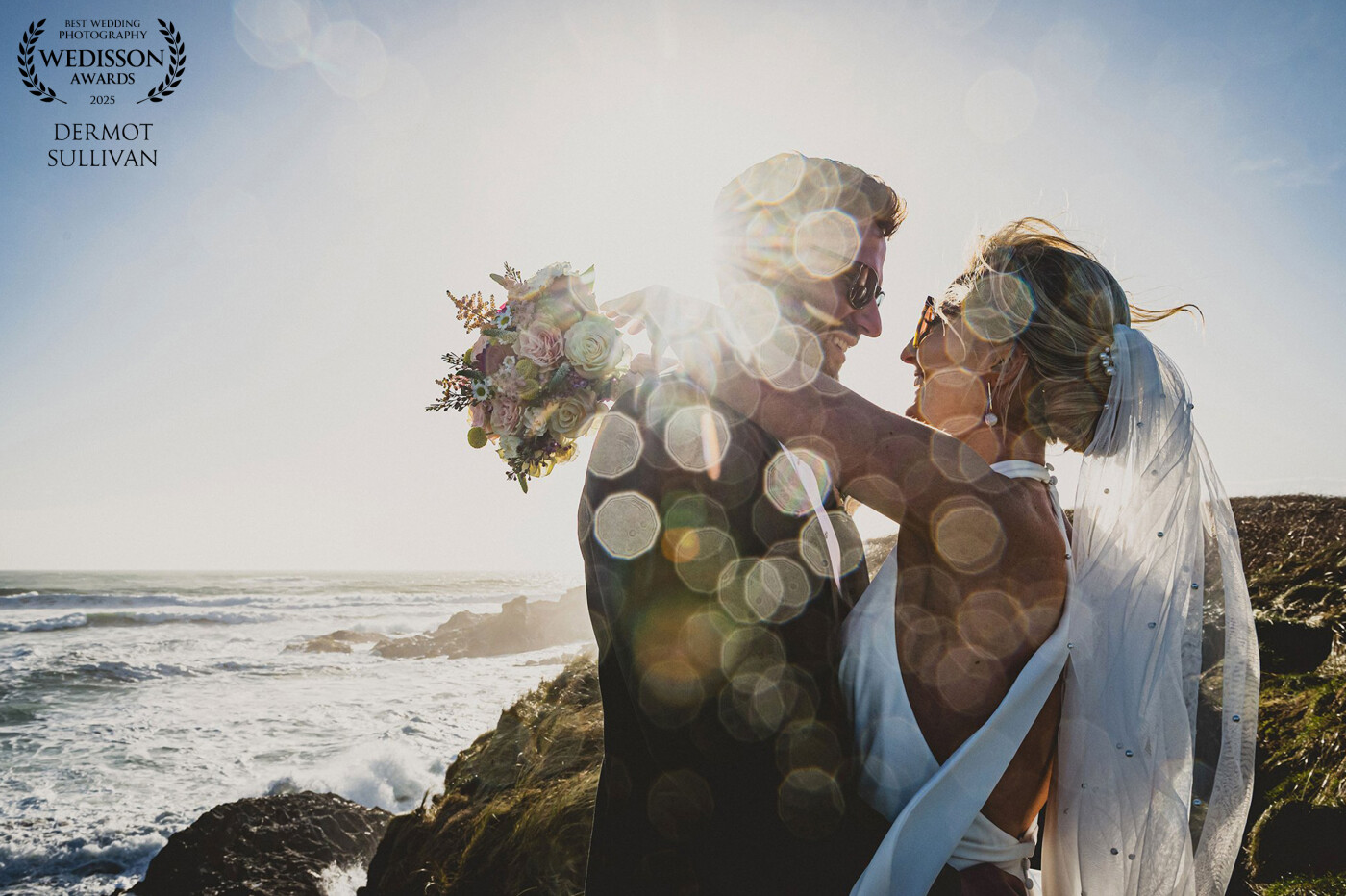 The brides in my two successful photographs in Collection 120 are sisters who were married a few months apart from each other. These are the 3rd and 4th sisters that I have photographed in their family! <br />
<br />
In this image, we went to a headland overlooking a beach in West Cork, the sun was shining strongly and I used a wide lens to capture the sunburst over the grooms shoulder while they were embracing.