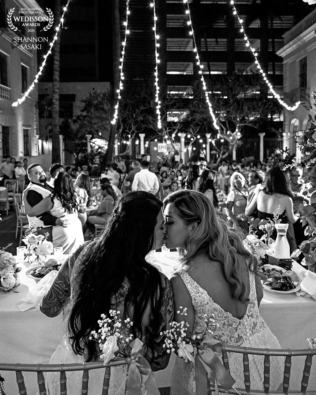 Two brides share a quiet kiss under a canopy of twinkling lights, surrounded by the joy and warmth of their loved onesโan unforgettable moment of love in the heart of celebration.